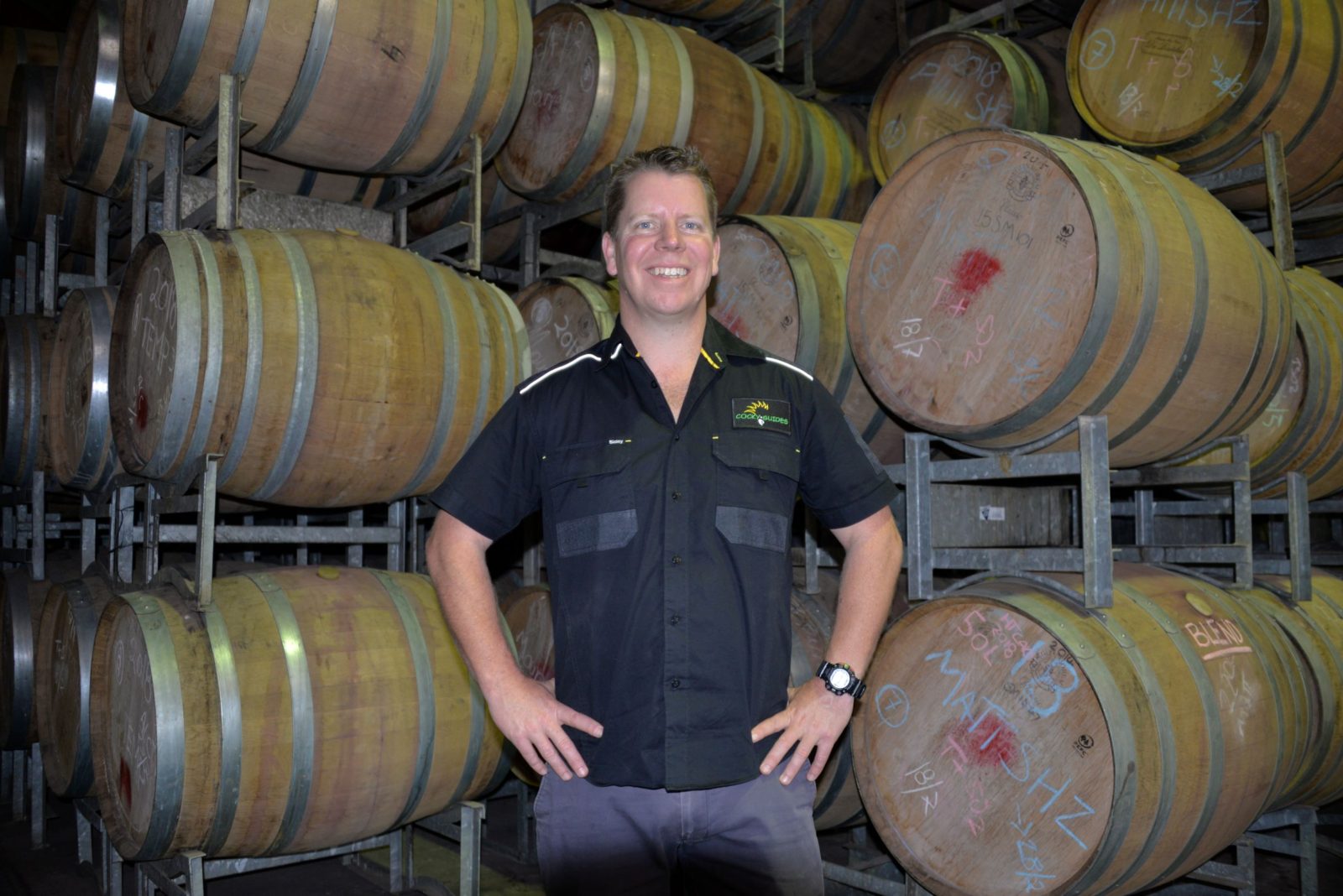 Cocky Guides Leader Buck standing in a wine storage area at a winery with oak barrels stacked behind him