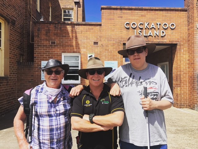 Cocky Guides Leader Buck between low vision travellers with white canes Paul and Sam at the entrance to Cockatoo Island in Sydney Harbour