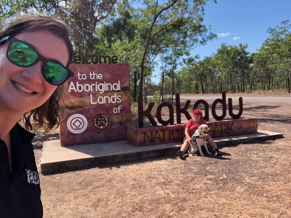 Selfie of trip leader Liz with Cocky Guides traveller Roz and Seeing Eye Ringo in the background seated by the entrance sign to Kakadu National Park