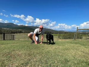 Trip Leader Dutchy crouched down next to black dog guide on lawn in the Victorian Alps on a sunny day