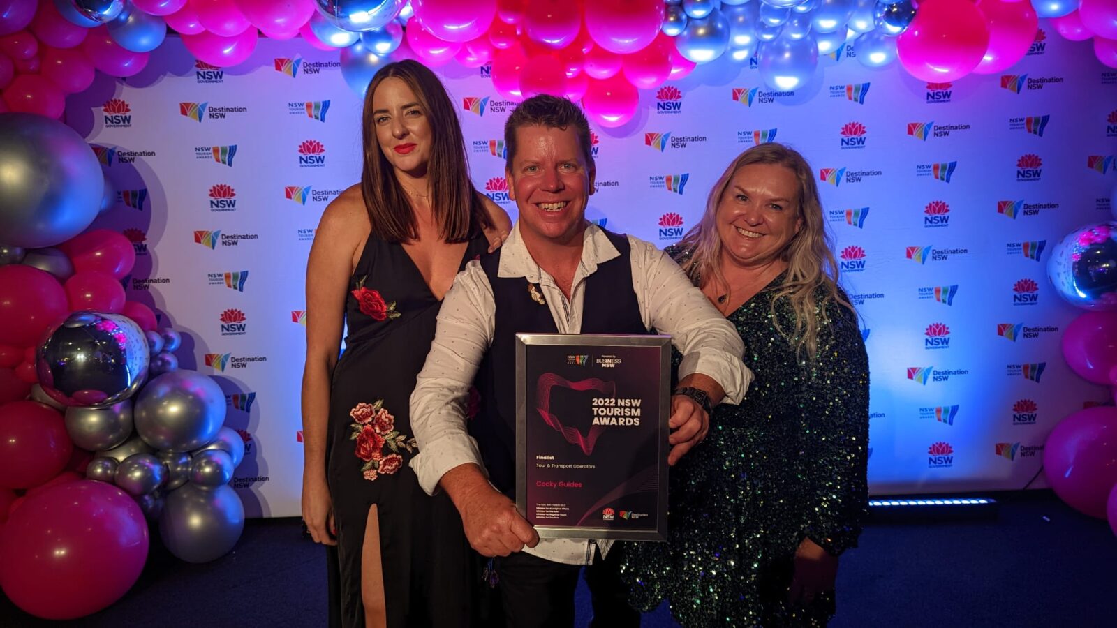 Cocky Guides Founder James (Buck) McFarlane holds the framed award with Trip Leaders Kellie and Donna on either side. They are indoors in a function room and stand in front of the event media wall with balloons around.