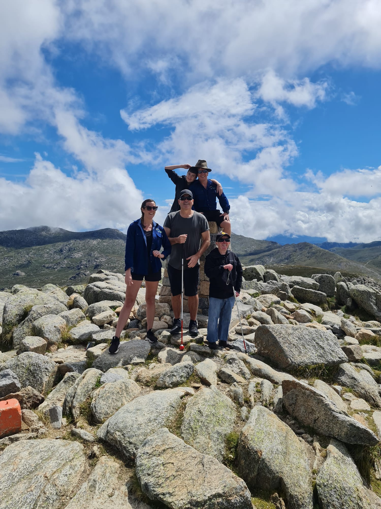 Four white cane users at the top of Mount Kosciuszko with two trip leaders on a sunny day