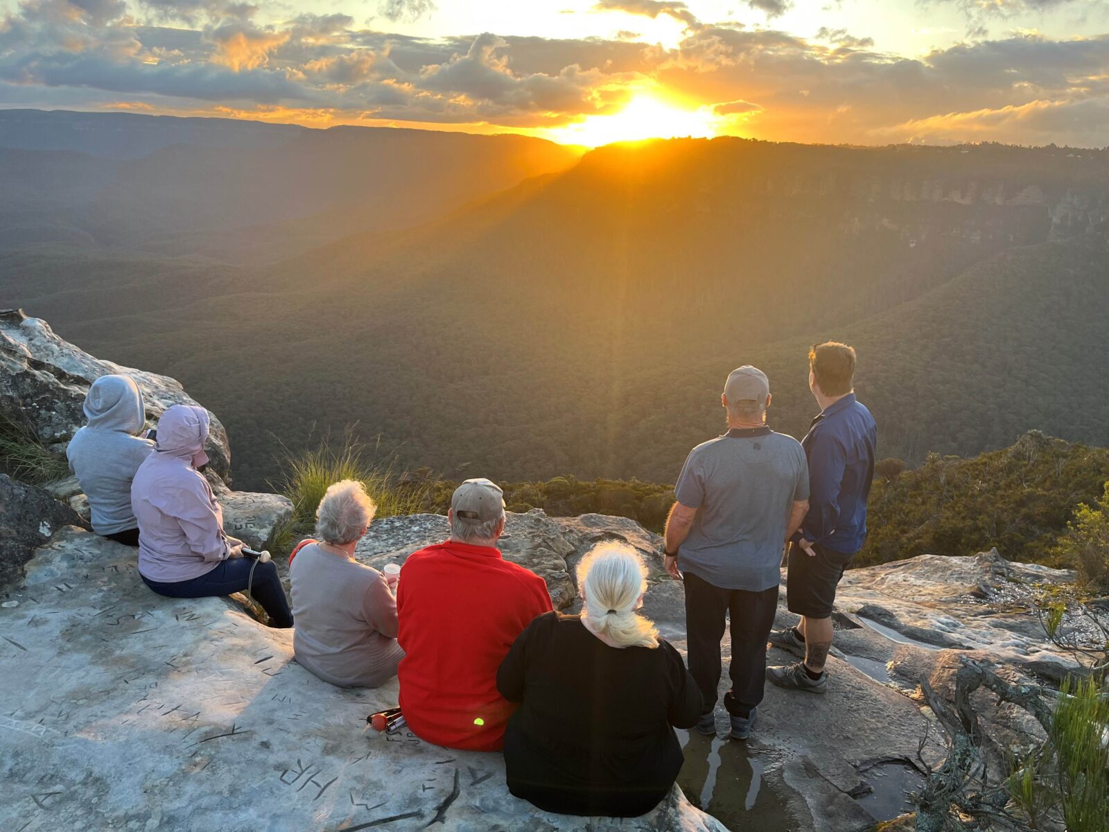 Five travellers seated on sandstone rock looking across Jamison Valley in the Blue Mountains at sunset. The trip leader and another travel is standing next to them on the right.